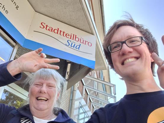 Zwei fröhlich grüßende Personen vor einem Schild auf dem "Stadtteilbüro Süd" steht. Die linke Person ist weiblich gelesen, die rechte männlich.