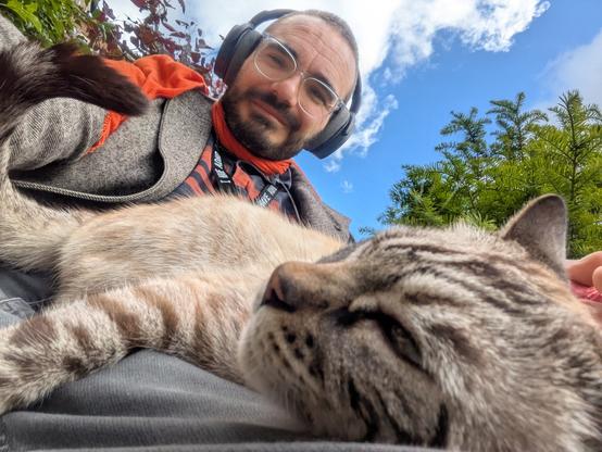 A selfie of Will, sitting down, taken from knee level. In the foreground, Tony, a Siamese/tabby cat  is lying down on Will's leg,  head resting sideways, looking pretty chill.