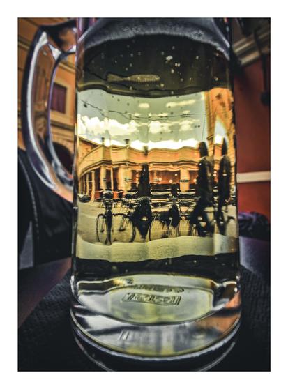 This photo is of the distorted reflection/refraction of a street in Bologna - through a large glass mug filled with a light-coloured beer. The reflection includes people cycling, and an arched building facade which are bent and warped by the glass and liquid inside. There is some slight bubbling and foam towards the top of the glass above the cyclists. Outside of the glass is not very in focus but you can make out that it is sitting on a black table and there are yellow and dark red buildings behind it.