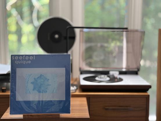 Seefeel - Quique LP cover.

Blue background, with a very light blue center with a 3-D spiked shape surrounded by light parentheses. Title and band upper left.

The black LP plays on a vintage, restored Bang & Olufsen Beogram 4000 linear-tracking turntable to the right.