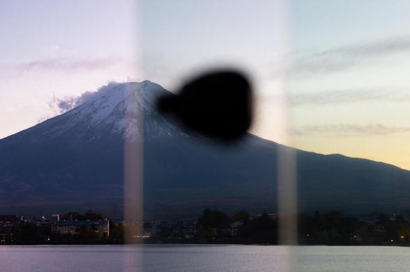 Mount Fuji with a snow-capped peak is visible in the background against a twilight sky. A large black silhouette obscures part of the mountain, situated in the center. In the foreground, there is a body of water with a shoreline featuring buildings and trees. The image is divided vertically by lighter bands on either side of the silhouette.