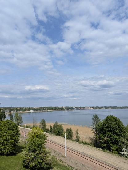 Spokojna tafla jeziora, na pierwszym planie tory kolejowe. W tle widać panoramę Giżycka.