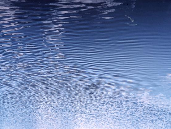 The surface of a body of water, under a partly cloudy sky. The water exhibits a deep blue hue, with lighter blue areas reflecting the sky and some darker, more opaque sections. The surface of the water is not perfectly still but has numerous small, rippling waves that create a textured pattern across the entire frame. 

To confuse the unimaginative, this photo has been rotated 180 degrees.