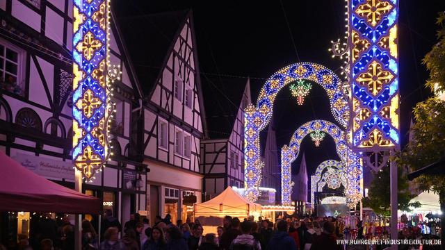 Festlich beleuchtete Fachwerkhäuser und bunt geschmückte Lichtbögen bei Nacht in der Innenstadt von Unna während des Un(n)a Festa Italiana. Zahlreiche Menschen schlendern durch die Straßen, über ihnen leuchten kunstvolle Lichtinstallationen in Blau, Gelb und Weiß.