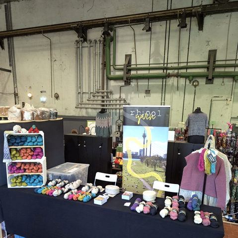 Ein schwarzer Ausstellungstisch auf dem verschiedenste farbige Wollsträge dekorativ drapiert sind. Im Hintergrund ein großes Banner von "rhool yarn". Noch weiter im Hintergrund sieht man die Wand einer Industriehalle, mit Rohren und Kabeln
