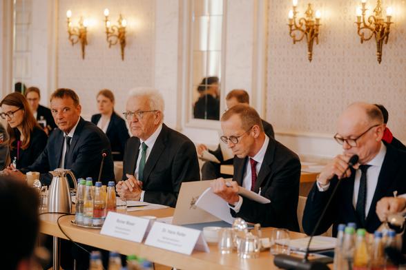 Fünf Personen in formeller Kleidung - darunter Ministerpräsident Winfried Kretschmann - sitzen an einem Konferenztisch mit Mikrofonen, Getränken und Namensschildern.
