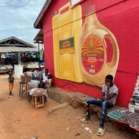 Scene from outside an market building in Uganda, with stalls selling goods. The side of the building is covered with a large hand-painted sign in bright reds, yellows, and greens, with realistic pictorials of two cooking oil containers branded 'Fortune'.