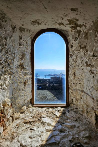 A tall, arched wooden window set within a rough, light-coloured stone wall of an old castle. Through the window, a bright blue sky meets a calm sea with a distant coastline. Below, a harbour is visible with boats and docks. Sunlight casts a distinct shadow on the stone floor in the foreground.