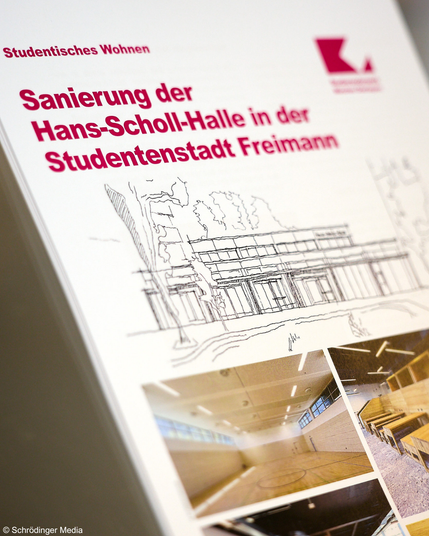 Foto, auf dem ein Papier zu sehen ist. Titel: Sanierung der Hans-Scholl-Halle in der Studentenstadt Freimann. Darunter eine Zeichnung eines Flachbaus sowie im Anschnitt ein paar Fotos.