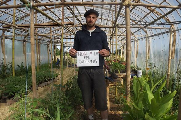 Un homme debout dans une serre de production en maraîchage bio, portant une feuille sur laquelle est écrit "NON À LA PPL DUPLOMB" 💀