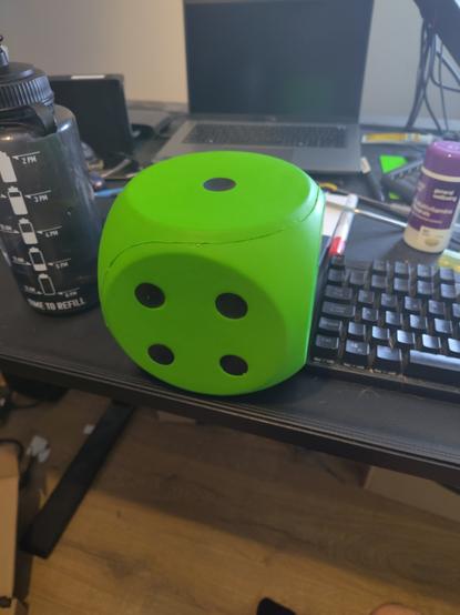 Photo of a messy black desk. In the background, a laptop can be seen with its screen off. In the foreground is a giant neon-green foam die, with black spots. The side facing the camera is showing 4, and the top is showing 1. The die is approximately 15cm to a side, and  is flanked by a 1L water bottle and a mechanical keyboard, both of which look somewhat smaller than you'd expect compared to the die