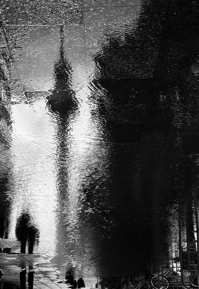 A black and white reflection on a wet surface shows distorted silhouettes of people, with an iconic tower visible in the background.