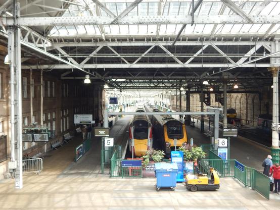 Two trains in yellow livery with red and grey accents in a train station.  Brick walls line the platform and a metal latticed roof covers the trains. Light comes in through a skylight.