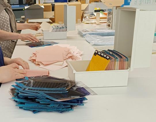 2 boîtes en plastique beige avec des intercalaires, ouvertes sur une table et vues en perspective. 2 collègues enlèvent les papiers qui protège chaque microfiche de taille rectangulaire et de couleur bleue. Ces microfiches reproduisent en petites images le Journal officiel et d'autres publications officielles. 
A l'arrière-plan, des boîtes d'archives grises et beiges.