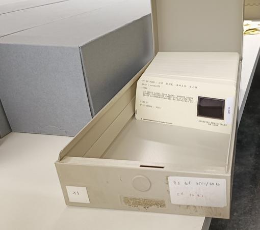 Sur une table claire, une boîte métallique longue et beige est ouverte. Elle contient des fiches cartonnées beige d'un format allongé, qui comprennent une petite reproduction sur film souple : il s'agit de "cartes à fenêtre". Le titre du document et sa cote sont dactylographiés.
A gauche, de grands cartons gris.
