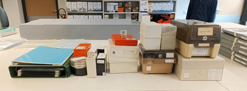 Sur une table claire, un ensemble de boîtes qui ont contenu des microfilms, microfiches et cartes à fenêtre. Il s'agit de garder pour mémoire un exemplaire de chaque conditionnement utilisé.