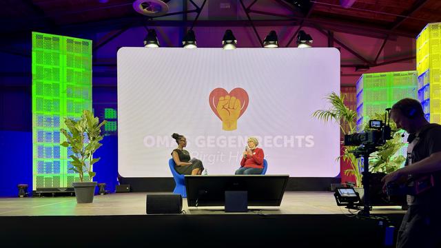 A stage with two women sitting in blue chairs engaged in conversation. The background features a large screen displaying the logo "OMAS GEGEN RECHTS" with a heart and raised fist symbol. There are green-lit decorations and a camera operator in the
