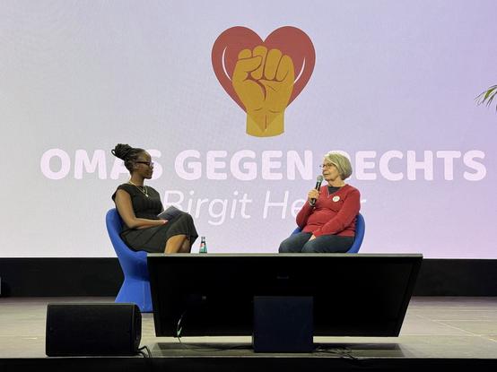 Two women are seated on a stage, engaged in conversation. One woman, wearing a black dress, is listening intently while holding notes. The other woman, dressed in a red sweater and glasses, is speaking into a microphone. A large backdrop features