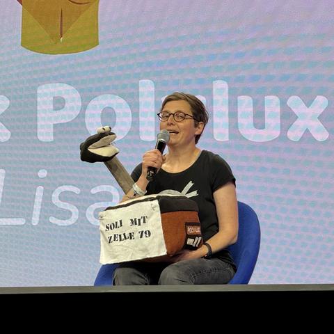 A woman is speaking into a microphone while holding a puppet. She is sitting on a chair in front of a colorful background with the words "Polylux" visible. The puppet appears to be part of an interactive presentation or performance.