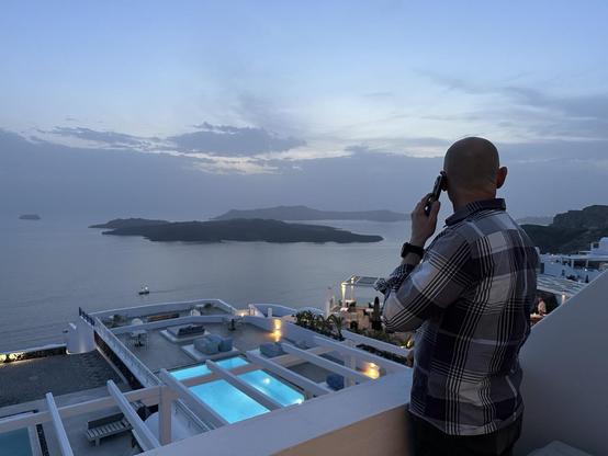 View from a balcony, overlooking the Aegean Sea. It’s dusk, and the sky above is mostly clear. The low clouds far away, where sky and sea meet, are almost the same blue-gray as the still waters. There is a small island in the sea about 2.5 kilometers from the vantage point, and a long way behind it resting on the horizon is the shade of another, larger island.

A man in a plaid shirt is talking on the phone while looking at the scene. The hotel is built in terraces, and on the terrace directly below we see an illuminated pool with lounging chairs around it. On a further terrace we can glean empty tables each with two chairs, like a small restaurant.