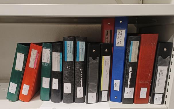 Une partie des classeurs d'origine posés sur une étagère métallique blanche, numérotés et étiquetés. Ce sont des classeurs en plastique de différents formats et couleurs.