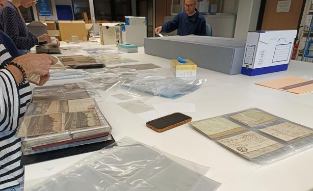 Sur une table blanche, une collègue qu'on voit en partie est en train de transférer les cartes postales dans des pochettes neuves.
On aperçoit 2 autres collègues.