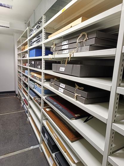 Dans un local de stockage peint au sol en noir et aux murs blancs, vue en perspective de rayonnages où sont rangés des boîtes et des documents de grandes dimensions.