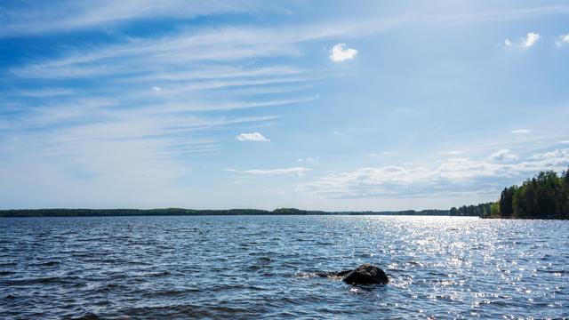 A vast expanse of water under a bright blue sky, with scattered clouds. The sunlight creates glittering reflections on the water's surface. A dark rock protrudes from the water in the foreground, and tree-lined shores are visible in the distance on the right side of the image.