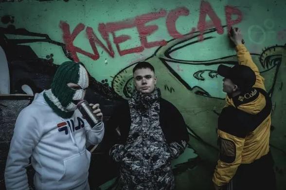 A publicity still of the band Kneecap standing in front of was graffiti bearing their name.
