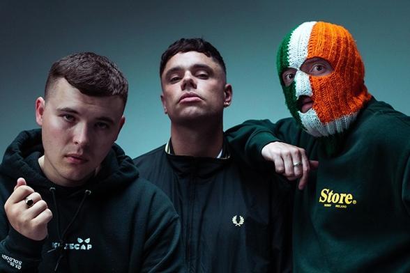 A publicity still that shows the members of the Irish Hip Hop band kneecap. All three are facing the camera. From left to right there is Mo Chara, Moglai Bap and DJ Provai.