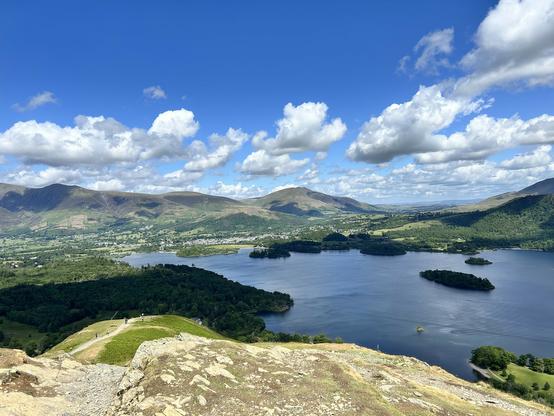 The image depicts a scenic landscape featuring a large lake surrounded by rolling hills and mountains. In the foreground, there is a rocky outcrop with patches of grass and a path leading down towards the lake. The lake is calm and reflects the blue sky, with several small islands covered in greenery scattered across its surface. The hills in the background are lush and green, with some areas appearing more barren. A small town or village is visible in the valley, with buildings clustered together. The sky is bright blue with scattered white clouds, suggesting a clear and sunny day. The overall scene is tranquil and picturesque, showcasing the natural beauty of the area.