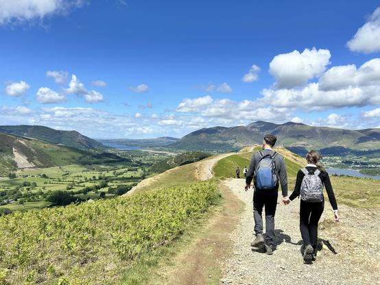 The image depicts a scenic outdoor hiking trail on a clear day. In the foreground, a man and a woman are walking hand in hand along a dirt path, both wearing backpacks and hiking attire. The man is wearing a gray jacket, black pants, and a blue backpack, while the woman is dressed in a black jacket, black pants, and a gray backpack. They are walking away from the camera, with the man slightly ahead of the woman.

The background features a vast landscape with rolling green hills and mountains under a bright blue sky with scattered white clouds. A valley with a body of water, possibly a lake, is visible in the distance, surrounded by lush green fields and patches of trees. The path they are on appears to be well-trodden, suggesting it is a popular hiking route. The overall scene conveys a sense of tranquility and natural beauty, with the hikers enjoying a leisurely walk in the countryside.