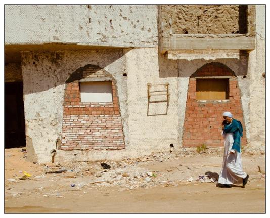 A man in white and blue traditional attire, talking on a mobile phone, walks past a dilapidated building with peeling white paint and exposed brickwork. The building has boarded and bricked-up windows, a dusty facade, and evident neglect, highlighted by scattered debris and litter in the foreground. The bright sunlight casts shadows on the uneven ground.