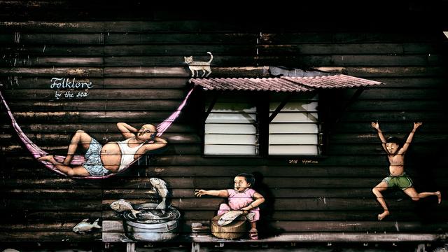 A mural on a wooden building shows a man relaxing in a pink hammock. Near him, a cat walks on a ledge. Below, a woman sits, holding a fish, and a pot with more fish is nearby. A boy in green shorts jumps joyfully beside them.