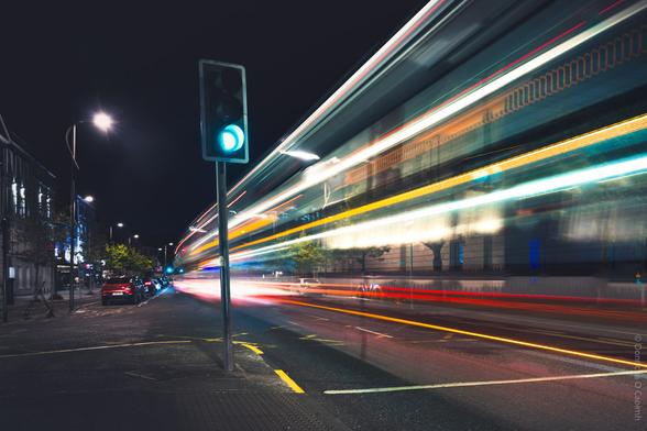 A long-exposure night photograph taken in Cork, Ireland, showing a city street with a green traffic light in the foreground and colourful light trails from passing vehicles, creating dynamic streaks of red, yellow, and white across the image; parked cars line the left side of the street, and buildings are visible under streetlights, with the sky dark above.