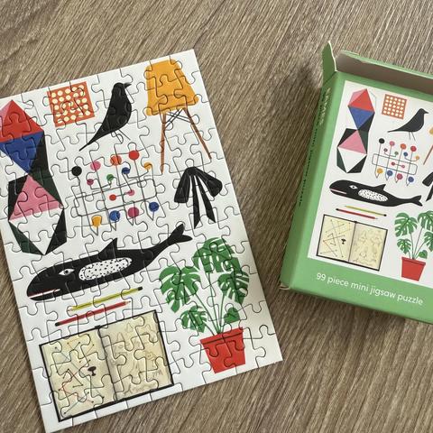 A completed 99-piece mini jigsaw puzzle featuring colorful, abstract shapes, a whale, birds, a potted plant, and a drawing. The puzzle is placed next to its green box, showcasing the design on the packaging.