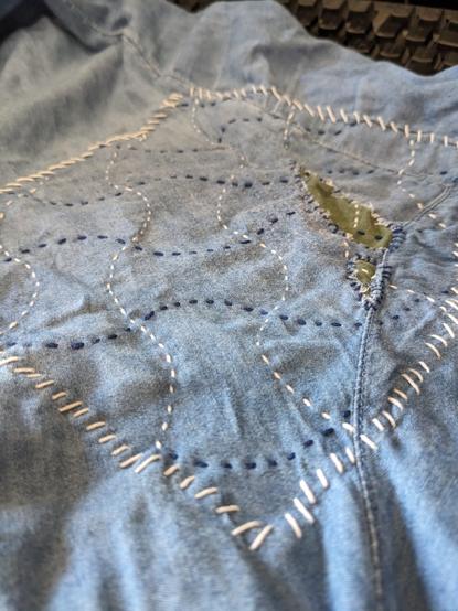 Close-up of a pair of pants with a large patch sewn on. It is done in a sashiko style, with this particular pattern being a sort of crisscross of wavy lines in white and blue thread.