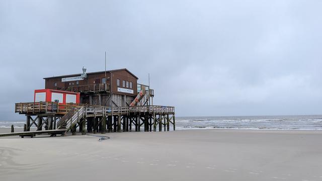 Ein Pfahlbau am Strand von St Peter Ording.
Das Haus steht auf hohen Pfählen, eine Treppe führt nach oben. Eine Veranda umgibt den Bau.