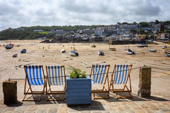 Four empty deck chairs on a harbour wall, taken from behind. They are made of blue and white striped canvas. There is a blue wooden box shaped flower pot in between the middle two, with some plants on. A stone pillar is at each end of the row of chairs. They overlook a harbour, empty of water, the sea at low tide. Boats rest on sand. A town is visible the other side of the harbor.