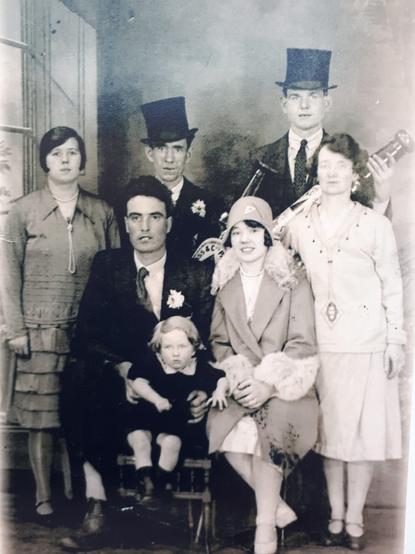 A black and white photo of a wedding group. At the front are seated a very tall groom and his bride wearing a very 20s outfit. The groom has a young child on his lap. At the sides stand two women. And behind are two tall men holding extremely large Guinness bottles.