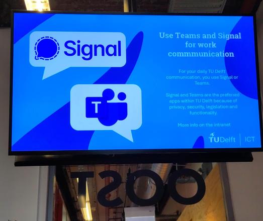 Foto van een beeldscherm op de gang bij faculteit bouwkunde waarop staat:
"use Teams and Signal for work communication" en tekst om verder op het intranet te kijken.
Voor Signal en Teams staan links blauwige logos,tekst staat rechts, achtergrond is TU-blauw.