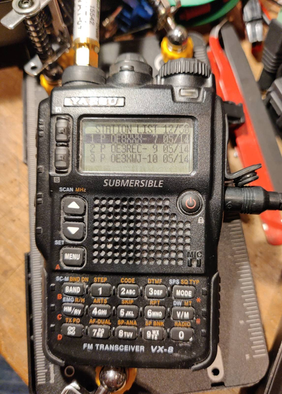 A picture of the again closed VX8 handheld radio with power attached, the radio is on and displays some old APRS stations in it's station list.