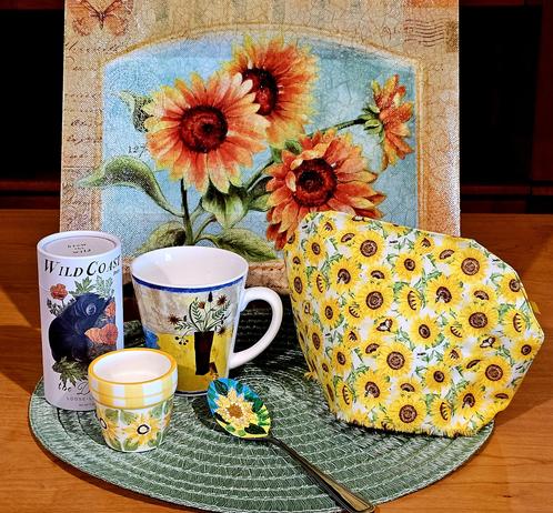 Tea tableau in bright sunny yellows and orange with light green and blue accents, of a sunflower theme. There are sunflowers on tea cozy cover, backdrop, china mug, miniature clay flowerpot, and a handpainted spoon. The tea is Wild Coast Brew loose-leaf herbal called Dreamer, in a white cylinder with artwork of a brown bear hugging California Poppies.