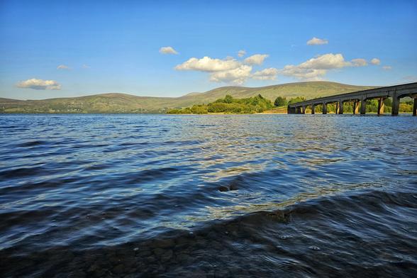 A wide shot of the Valleymount Bridge spanning a calm, dark blue lake under a bright blue sky with fluffy white clouds. In the foreground, the rippling water of the lake fills the bottom half of the frame, showing some of the rocky bed beneath the surface. The multi-arched concrete bridge extends from the right side of the image towards the centre, connecting to a distant treed shore. Rolling green hills rise in the background under the clear sky.