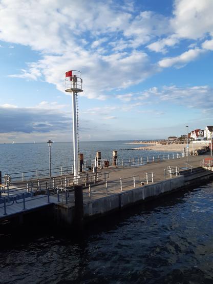 Blick auf einen Anlegesteg. Im Vordergrund ein Signalmast, im Hintergrund links Wasser, rechts Strand und weiße Häuser mit roten Ziegedächern. Himmel mit weißen Wolken und Wölkchen.