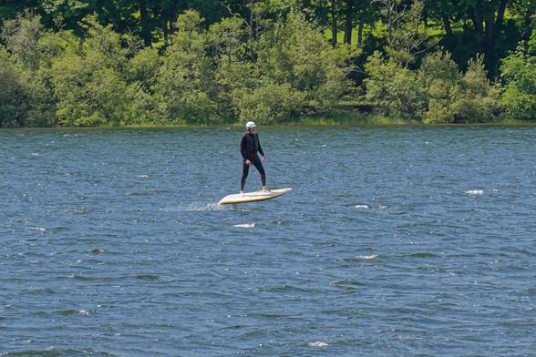 A long distance (and so slightly blurry) photograph of a man in a wetsuit standing on what looks like a short paddleboard. However looking closely it can be seen that the board is entirely out of the water and there is just a small vertical pipe sticking down into the water, which is where all the crazy clever shit happens.