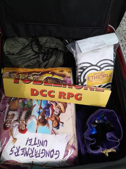 Photo of an open suitcase containing: a dice bag, a folder with "Gongfarmers Unite" on the front, a Toblerone chocolate bar with "DCC RPG" printed on the side, a bag of Ethiopian coffee. And a rain jacket (I am going to the UK after all).