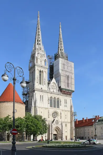 The image depicts a tall, architectural structure featuring two prominent spires. It seems to be a cathedral or a large church, characterized by its ornate façade and intricate details. There is scaffolding on part of the building, suggesting that restoration or maintenance work is taking place. In the surrounding area, there are trees, a circular flowerbed, and a street sign, indicating an urban setting. The sky is clear and blue, contributing to a vibrant atmosphere.

Image Credits: Wikimedia / Bernard Gagnon / CC BY-SA 4.0