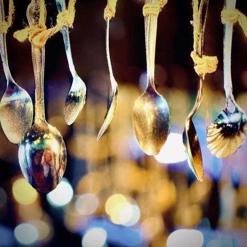Individual metal spoons hanging from coarse rope in focus in the foreground shine and reflect various amber and blue hues and sparkles out of focus in the background.