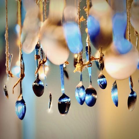 Individual metal spoons hanging from coarse rope in focus in the background shine and reflect various amber and blue hues and sparkles out of focus in the foreground.